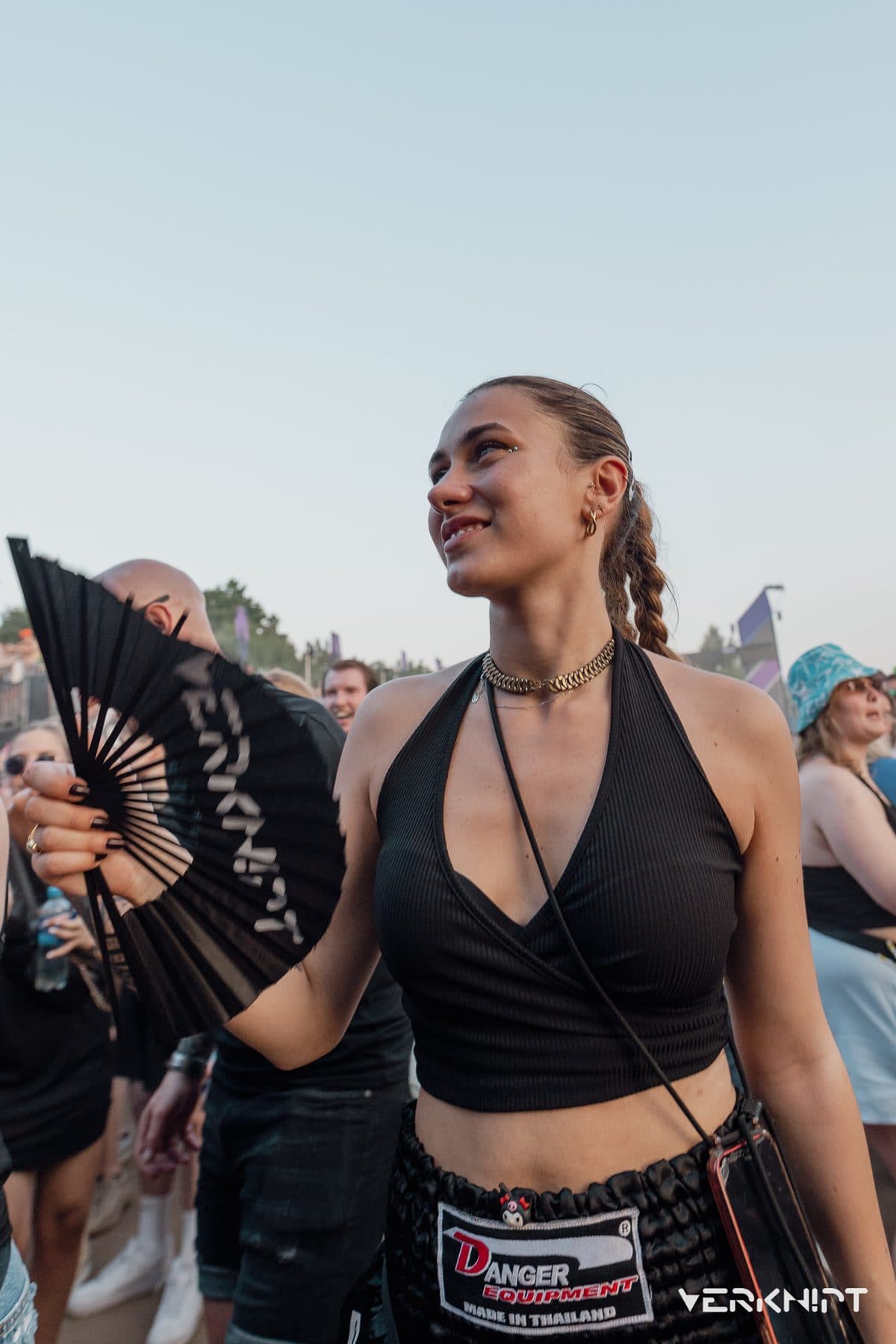 A festivalgoer holding an open black hand fan, walking through a daytime crowd against a pale sky.