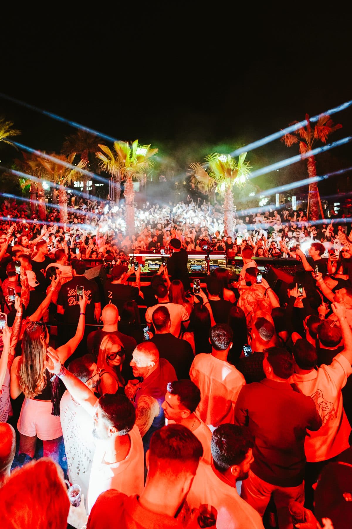 A packed open-air crowd under red light, laser beams crossing above the palm trees.