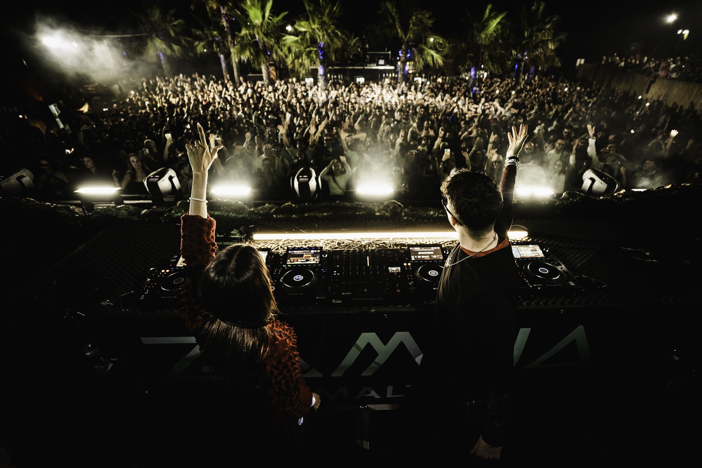 Two DJs seen from behind at the decks, arms raised, facing a packed night crowd under floodlit palm trees.