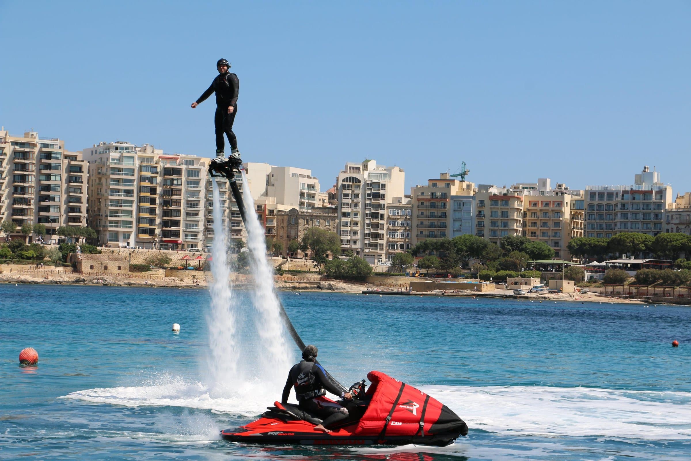 A flyboarder hovering above the water on a jet of spray, with the Sliema seafront behind.