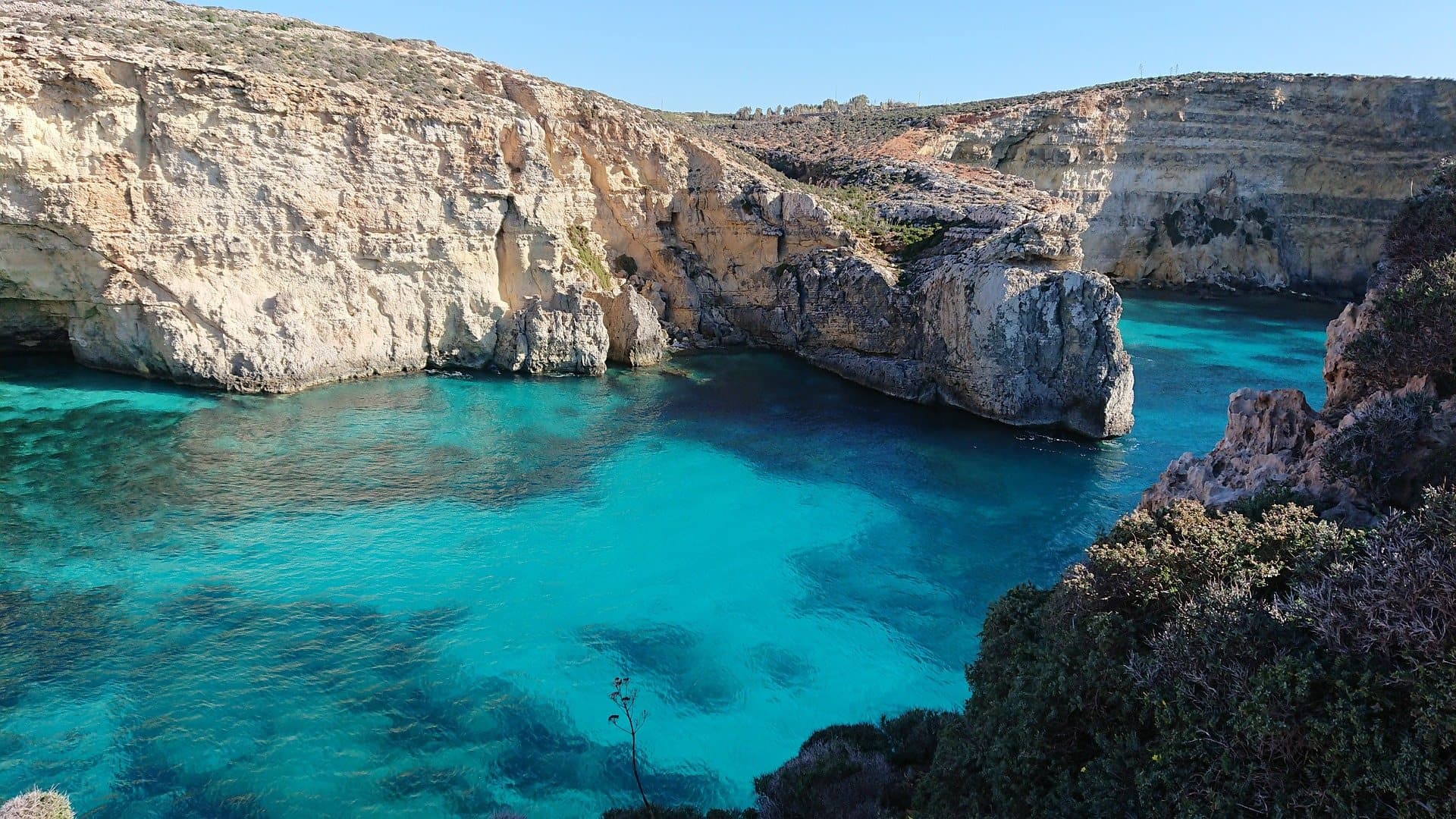 The Blue Lagoon at Comino from above, a pale limestone cove opening into water that runs from clear shallows to deep blue.