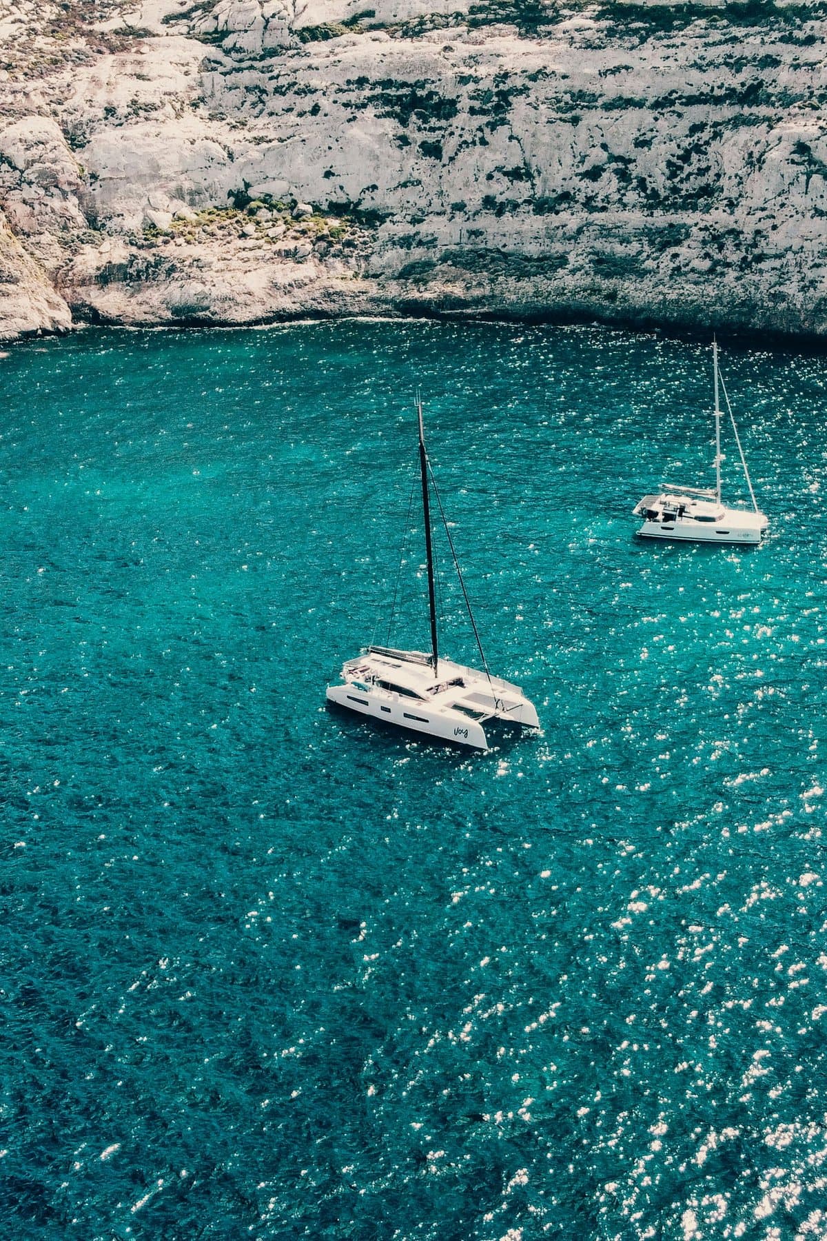 Two catamarans moored on flat turquoise water beneath Malta's pale limestone cliffs.