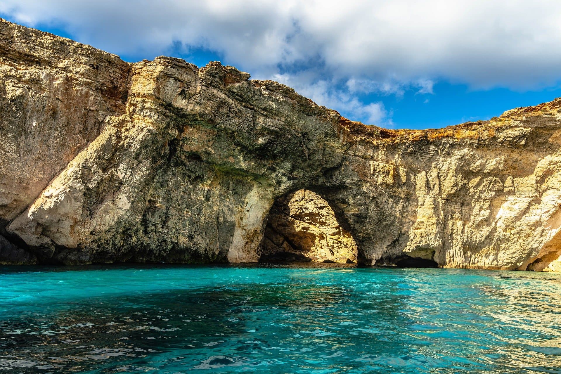 A natural limestone arch rising out of turquoise water off Comino, under an open blue sky.