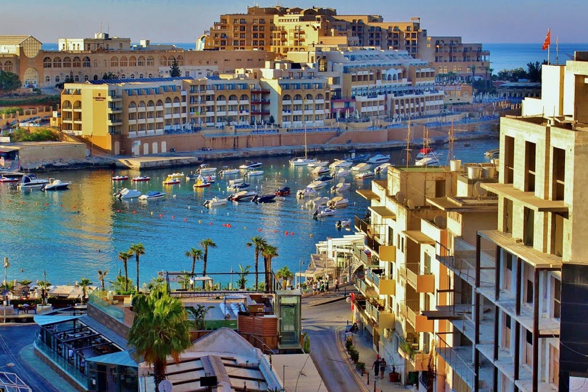 St Julian's bay at golden hour, moored boats on still water with palms and terraces along the shore.